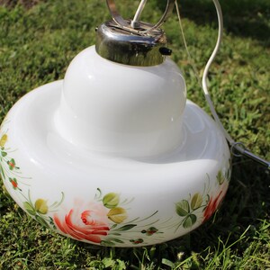 以下が含まれることがあります： 花柄の白いガラス製のペンダントライト。照明器具は、丸いボウル型のシェードに、さらに小さい丸いボウル型のシェードが重ねられています。花柄は、赤いバラ、緑の葉、黄色のアクセントが特徴です。