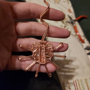 May include: A copper wire sculpture of an alien creature with a long, thin tail. The creature has a segmented body and eight legs.
