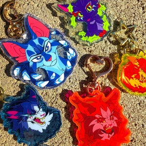 May include: A collection of colorful cat-themed keychains. The keychains feature various cat illustrations in shades of blue, purple, red, and orange. Each keychain has a gold-colored clasp. The keychains are made of clear acrylic.