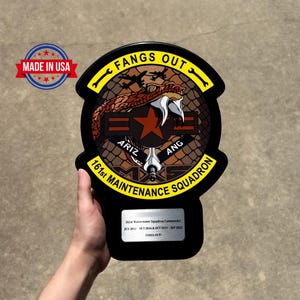 May include: A black plaque with a military emblem featuring a snake, star, and text "FANGS OUT." The plaque also includes the text "161st MAINTENANCE SQUADRON" and "MADE IN USA."