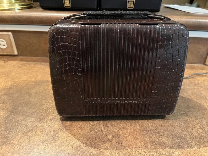 May include: A brown vintage Emerson Radio & Phonograph Corporation Model 111 radio with a crocodile-embossed leatherette case and a metal handle.