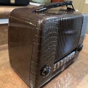 May include: A brown vintage portable radio with a crocodile-embossed design and a black leather carrying handle. The radio has a metal grille and a dial with gold lettering.