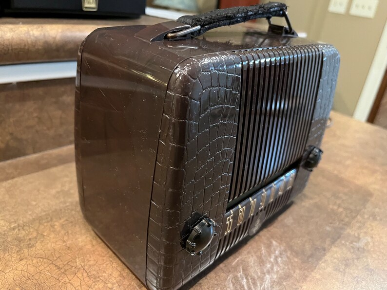 May include: A brown vintage portable radio with a crocodile-embossed design and a black leather carrying handle. The radio has a metal grille and a dial with gold lettering.