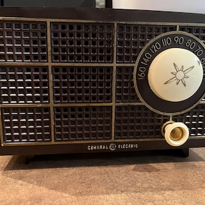 May include: A vintage brown General Electric radio with a white dial and a gold knob. The dial has numbers from 55 to 160.