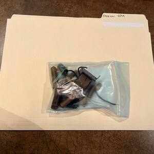 May include: A clear plastic bag containing a variety of electronic components, including resistors, capacitors, and a small black circuit board. The bag is sitting on a beige file folder labeled "Emerson 55YA".