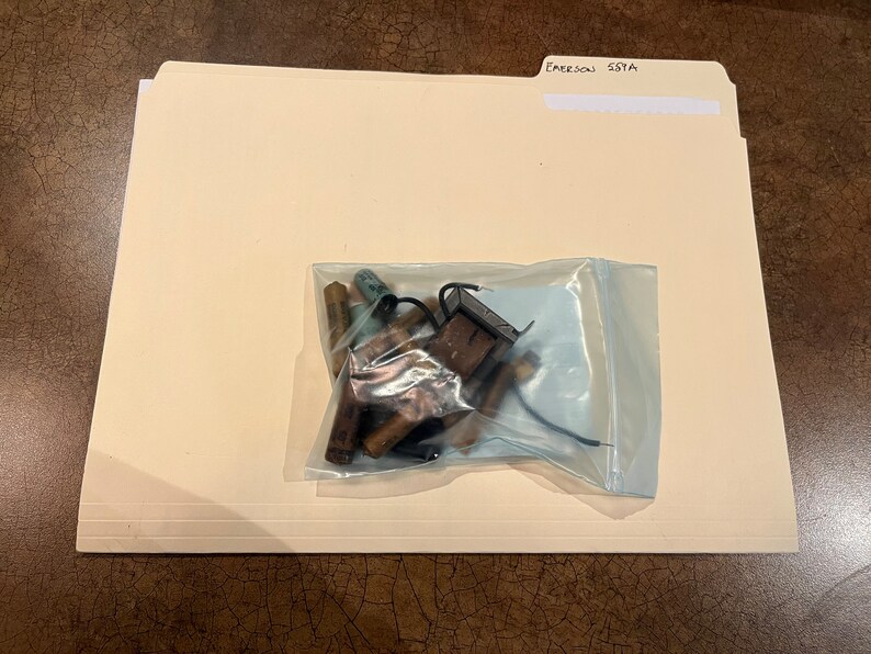 May include: A clear plastic bag containing a variety of electronic components, including resistors, capacitors, and a small black circuit board. The bag is sitting on a beige file folder labeled "Emerson 55YA".