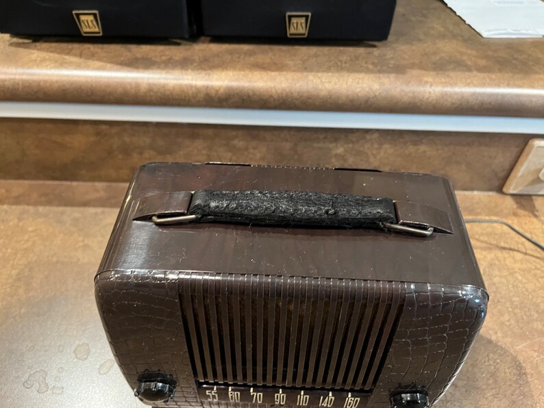 May include: A brown vintage radio with a black leather handle and a crocodile-embossed design. The radio has a dial with numbers from 55 to 180.