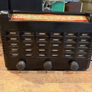 May include: A vintage black radio with a red and gold frequency dial. The radio has three knobs and a speaker grill with a geometric pattern. The radio has a cord and a white and black plug.