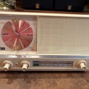 May include: Vintage cream-colored radio with a clock face on the left and a speaker on the right. The clock has a pink face with black numbers and hands. The radio has knobs for volume and tuning.