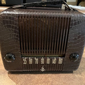 May include: A vintage brown radio with a crocodile-embossed design. The radio has a black dial with white numbers and a black knob on each side. The radio has a black handle on the top.