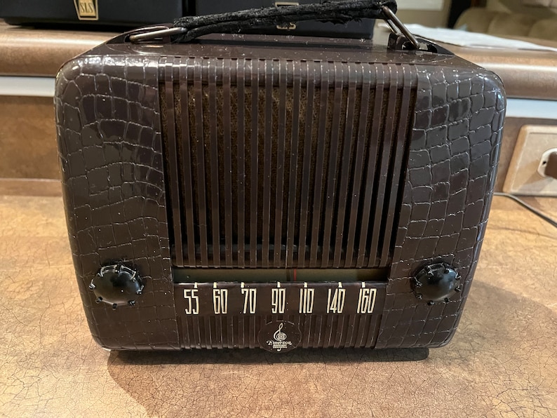 May include: A vintage brown radio with a crocodile-embossed design. The radio has a black dial with white numbers and a black knob on each side. The radio has a black handle on the top.