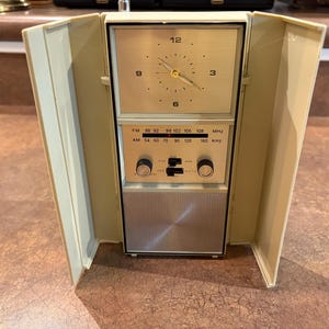 May include: Vintage cream-colored clock radio with a fold-out design. The clock face is gold with black numerals and hands. Below the clock are radio controls and a speaker. The radio has FM and AM frequency options.