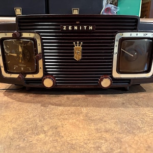 May include: A vintage Zenith radio with a dark brown finish. The radio features two square clocks on either side of the speaker grill. The Zenith logo is visible above the grill. The clocks have gold-colored frames and dark faces.