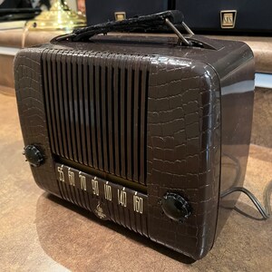 May include: A brown vintage radio with a crocodile-embossed design. The radio has a dial with numbers from 55 to 160 and a black knob on the right side. The radio has a black handle on the top.