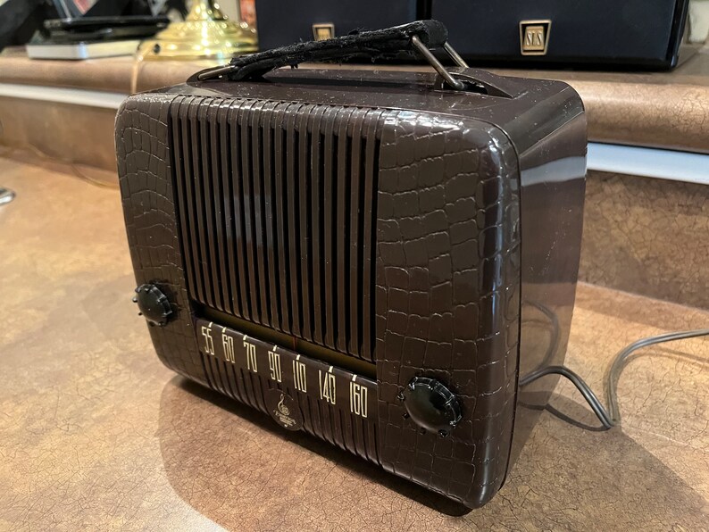 May include: A brown vintage radio with a crocodile-embossed design. The radio has a dial with numbers from 55 to 160 and a black knob on the right side. The radio has a black handle on the top.