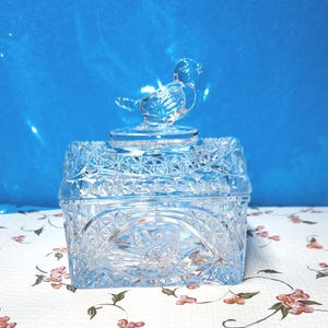 May include: A clear, square-shaped glass trinket box with a detailed, cut-glass design. The lid is topped with a small, transparent bird figurine. The box sits on a floral patterned surface against a blue background.
