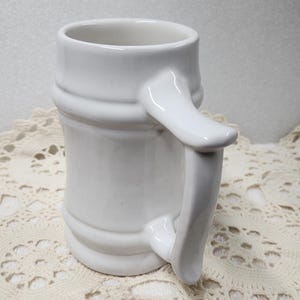 May include: A white ceramic mug with a handle. The mug has a cylindrical shape with two horizontal ridges. The handle is curved and attached to the side of the mug. The mug is sitting on a cream-colored, crocheted doily.
