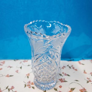 May include: A clear, cut-glass vase with a flared top and a diamond pattern on the lower half. The rim has a scalloped edge. The vase sits on a floral patterned surface against a blue background.