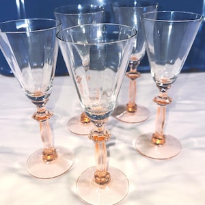 May include: Set of five clear wine glasses with a pink stem and base. The glasses have a triangular bowl shape and a decorative design on the stem. The glasses are arranged on a white surface.