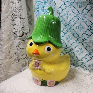 May include: A yellow ceramic chick figurine wearing a green leaf-shaped hat. The chick has a bright orange beak, black eyes, and is holding a small pink flower. Additional pink flowers and green leaves are painted around the base of the chick.