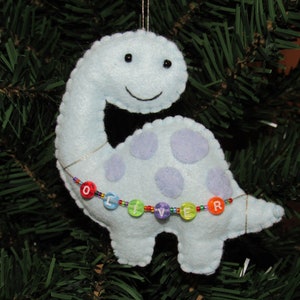 May include: A light blue felt dinosaur ornament with purple spots and a beaded necklace that spells "Oliver".