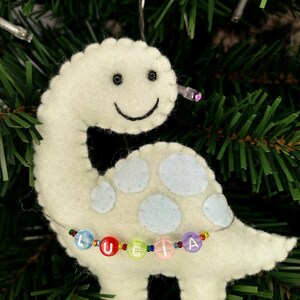 May include: A light green felt dinosaur ornament with a smiling face and blue spots. The dinosaur has a beaded necklace with the name "Lucia" spelled out in colorful beads.
