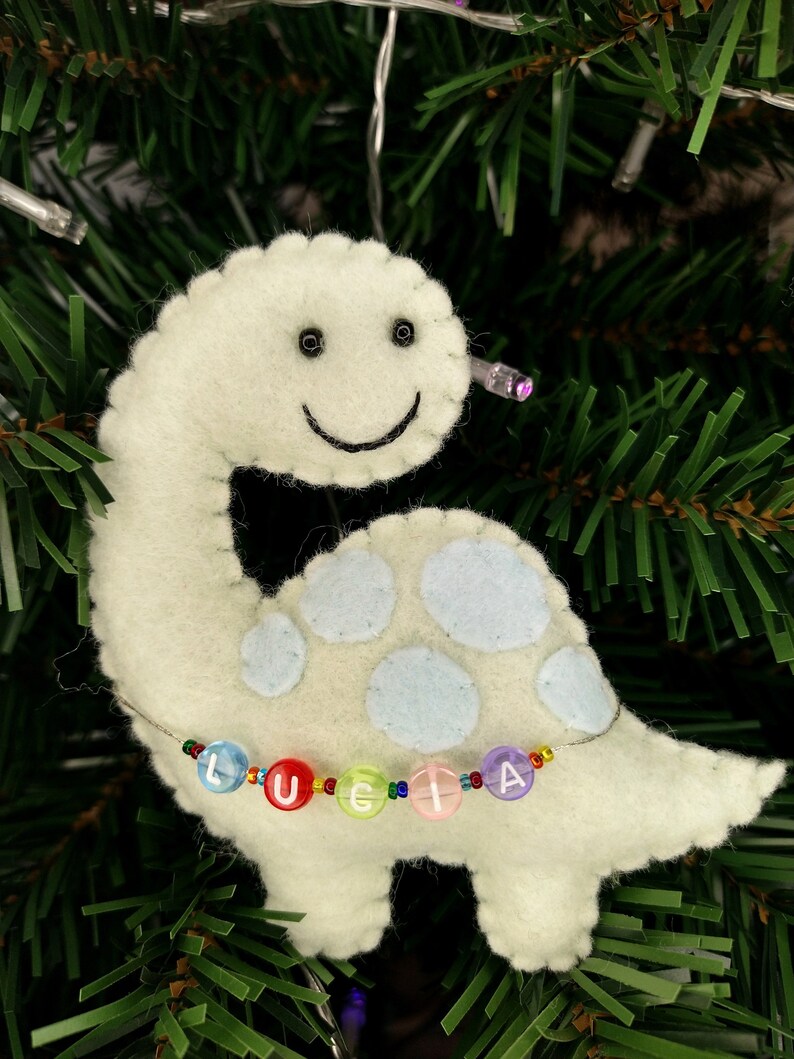 May include: A light green felt dinosaur ornament with a smiling face and blue spots. The dinosaur has a beaded necklace with the name "Lucia" spelled out in colorful beads.