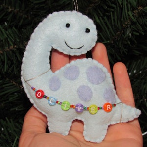 May include: A light blue felt dinosaur ornament with purple polka dots and a beaded necklace spelling "OLIVER".