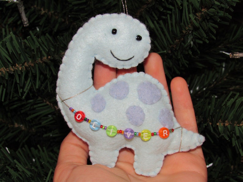 May include: A light blue felt dinosaur ornament with purple polka dots and a beaded necklace spelling "OLIVER".