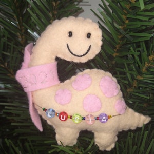 May include: A beige felt dinosaur ornament with pink polka dots and a pink ribbon. The dinosaur has a smiling face and the ribbon has the year 2021 embroidered on it. The dinosaur is holding a beaded string with the name "Lucia" spelled out in colorful beads.