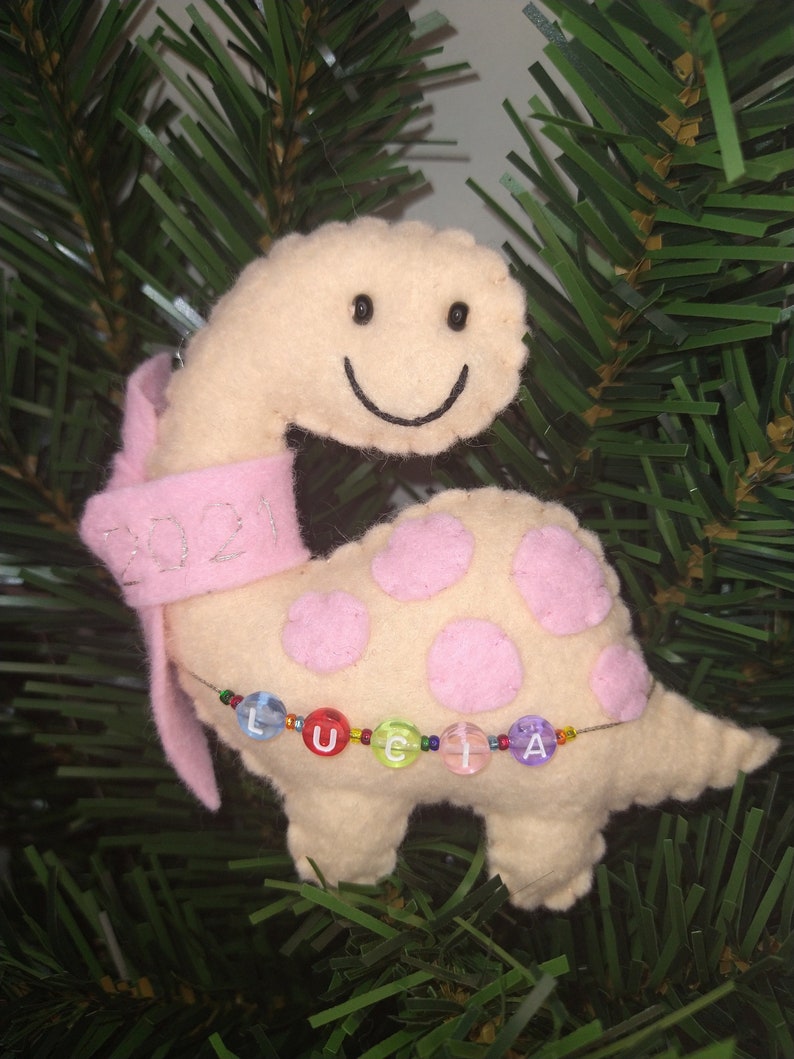 May include: A beige felt dinosaur ornament with pink polka dots and a pink ribbon. The dinosaur has a smiling face and the ribbon has the year 2021 embroidered on it. The dinosaur is holding a beaded string with the name "Lucia" spelled out in colorful beads.