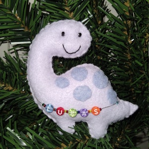 May include: A white plush dinosaur ornament with blue spots and a smiling face. The dinosaur has a beaded necklace with the name "LUCAS" spelled out in colorful beads.