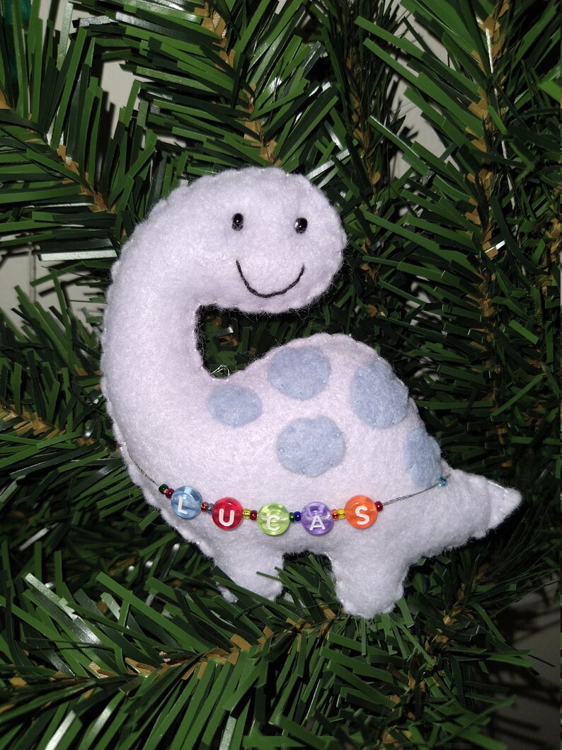 May include: A white plush dinosaur ornament with blue spots and a smiling face. The dinosaur has a beaded necklace with the name "LUCAS" spelled out in colorful beads.