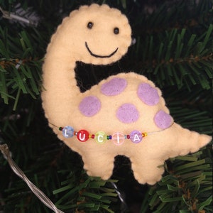 May include: A yellow felt dinosaur ornament with purple polka dots and a beaded name "LUCIA" hanging on a green Christmas tree.