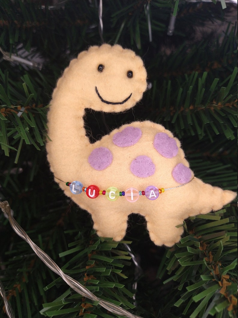 May include: A yellow felt dinosaur ornament with purple polka dots and a beaded name "LUCIA" hanging on a green Christmas tree.