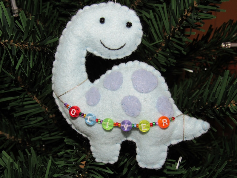 May include: A light blue felt dinosaur ornament with purple spots and a beaded necklace spelling "Oliver".