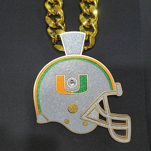 May include: A gold chain necklace with a silver and green glitter football helmet pendant. The helmet has a large letter "U" on the front with a diamond-shaped rhinestone in the center.