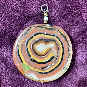 Könnte beinhalten: Ein runder, mehrfarbiger Anhänger mit einem Spiralmuster in Braun-, Gold-, Schwarz- und Weißtönen. Der Anhänger ist an einer silbernen Kette mit einer weißen Perle befestigt.