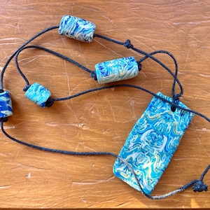 Könnte beinhalten: Eine blaue und weiße marmorierte Halskette mit einem rechteckigen Anhänger. Die Halskette besteht aus schwarzem Band und hat vier blaue und weiße marmorierte Perlen.