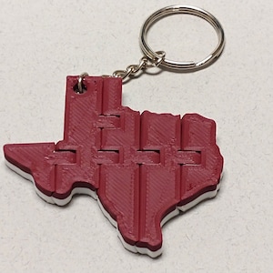 May include: A red 3D printed Texas state keychain with a silver key ring. The keychain is made of a textured plastic and has a white outline.