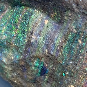 Peut inclure: Gros plan sur une roche rugueuse de couleur gris foncé avec des inclusions d'opale irisées vertes, bleues et violettes.