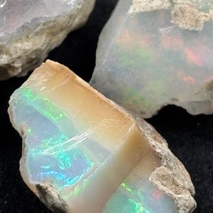 以下が含まれることがあります： いくつかの粗いオパール石のクローズアップ。オパールは、虹色の青、緑、赤の閃光を含むさまざまな色を示し、ニュートラルで明るい色の背景に設定されています。石は生の自然な外観をしています。