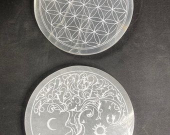 May include: Two white, round, selenite charging plates with etched designs. The top plate features a flower of life design. The bottom plate features a tree of life design with a sun and moon.