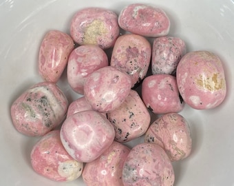 May include: A collection of pink and white tumbled stones with black and brown markings. The stones are smooth and polished, and they are arranged in a white bowl.