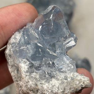 May include: A close-up of a celestite crystal cluster. The crystal is a pale blue colour with sharp, geometric formations. The base is a rough, grey-white rock, contrasting with the translucent crystal.