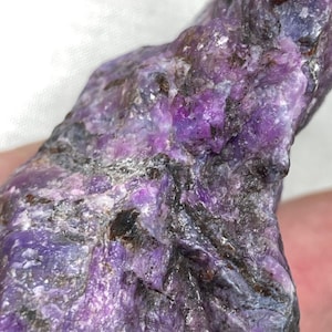 May include: Close-up of a raw, natural charoite gemstone. The stone displays a mix of vibrant purple hues, with areas of deep violet and lighter lavender. Darker, almost black, inclusions add contrast to the crystalline structure.