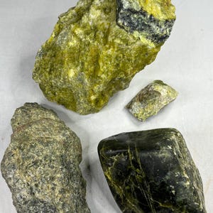 Pode incluir: Quatro espécimes minerais brutos em tons de verde e amarelo. A peça maior é um amarelo-esverdeado vibrante com inclusões escuras. Outras peças variam do azeitona ao verde escuro, mostrando texturas naturais.