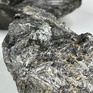 Puede incluir: Primer plano de una roca gris oscura con una superficie rugosa y texturizada. La roca tiene un brillo plateado y parece estar hecha de muchos cristales pequeños y delgados.