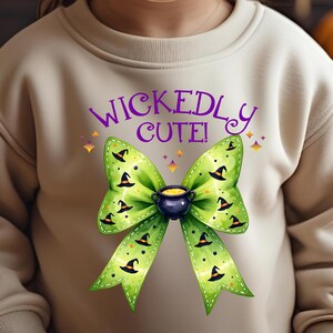 Pode incluir: Camisola bege com um laço verde com chapéus de bruxa e um caldeirão. O texto "WICKEDLY CUTE!" é roxo. Perfeito para o Halloween ou um evento temático assustador.