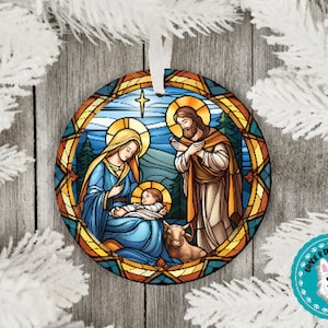 May include: A round Christmas ornament with a stained glass design depicting the Nativity scene. Mary, Joseph, and baby Jesus are surrounded by a golden halo and a star. The background is a blue sky with clouds.